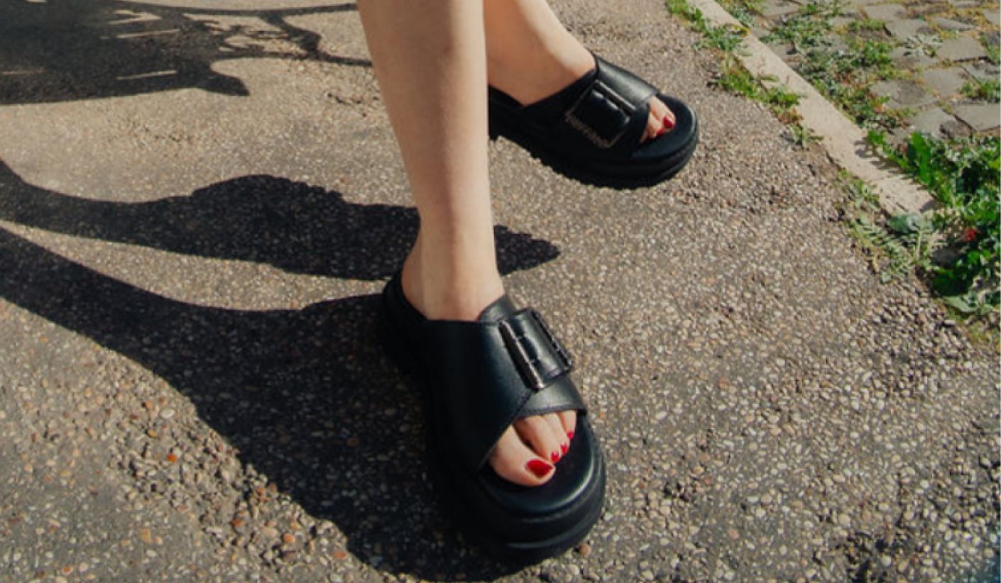 Close-up of black chunky slide sandals worn outdoors on asphalt pavement, featuring a wide strap and platform sole.