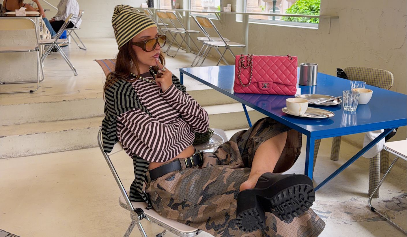 Close-up of a model seated in a café setting, wearing striped layers and chunky black slides, with a pink quilted handbag on the table.