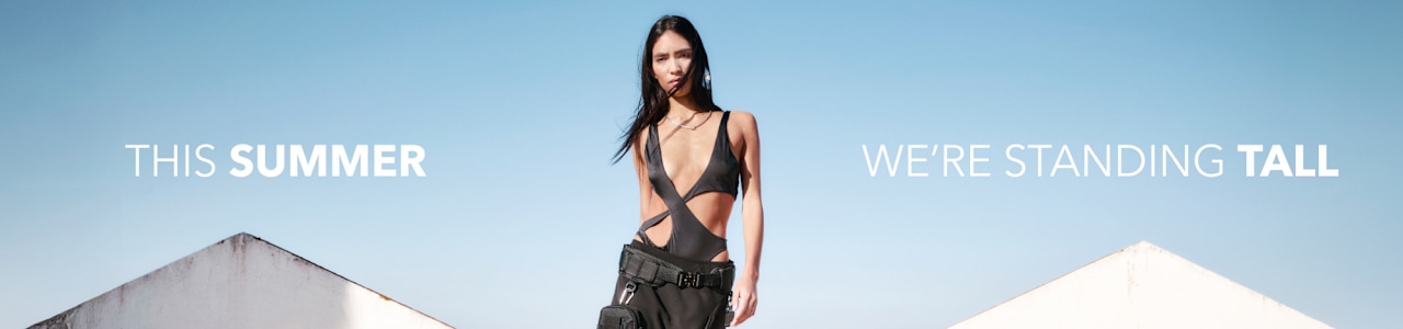 Model walking outdoors in a black cut‑out outfit and chunky shoes, with “THIS SUMMER WE’RE STANDING TALL” text overlay.