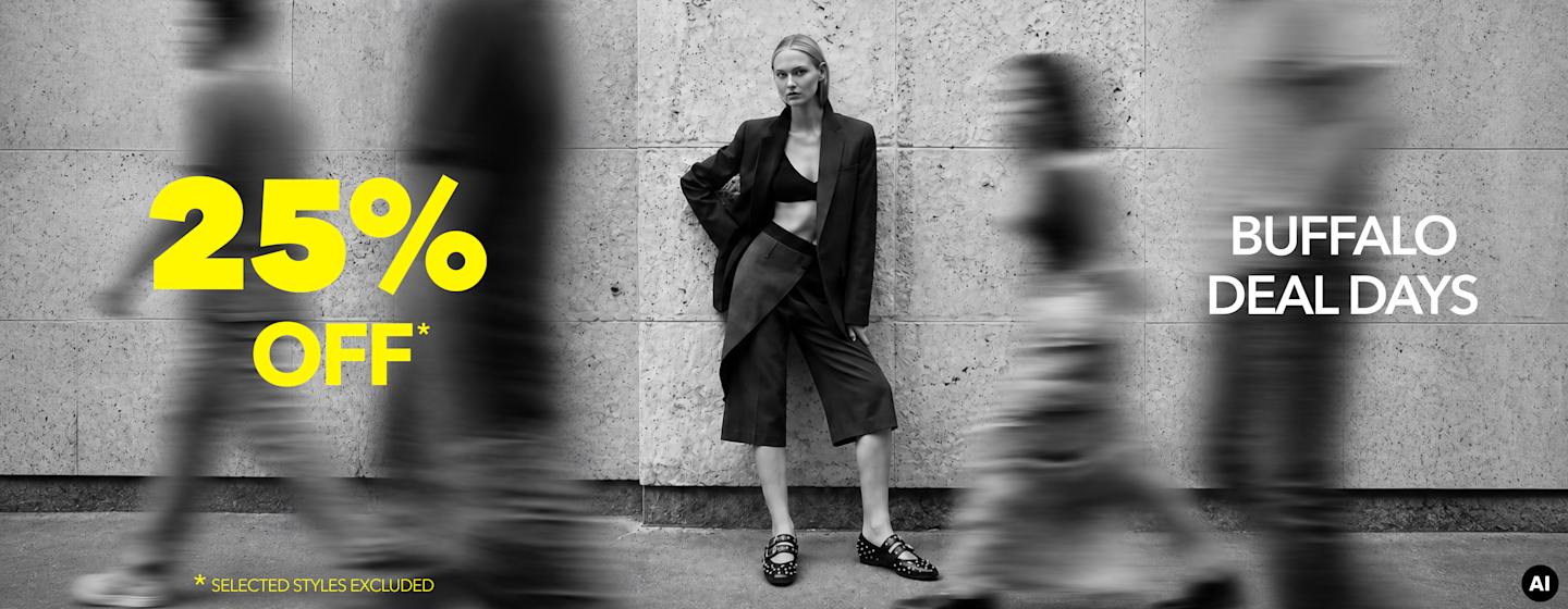 Person in a tailored black outfit and studded loafers standing against a stone wall, with blurred pedestrians and promotional sale text in the foreground.