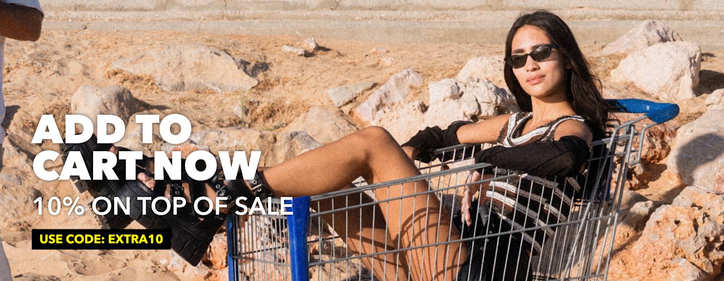 Fashion model sitting in a shopping cart on a rocky outdoor landscape beside a sale promotion banner.