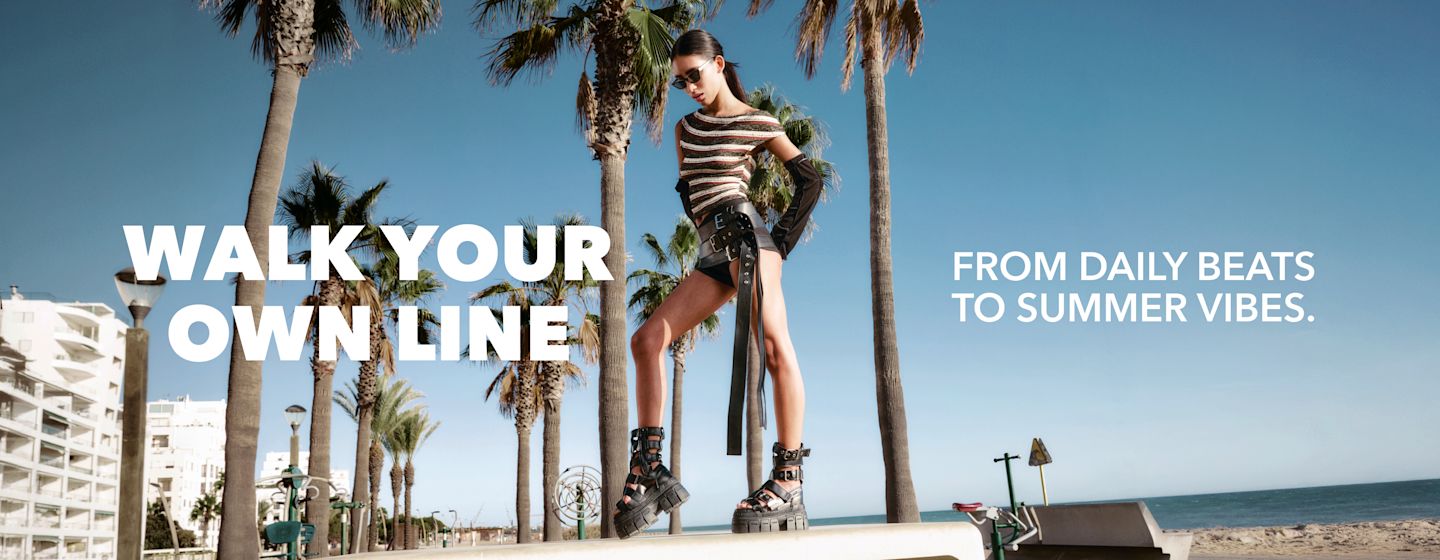 Fashion model in edgy outfit and platform sandals standing on a palm-lined beach promenade with bold “Walk Your Own Line” slogan.