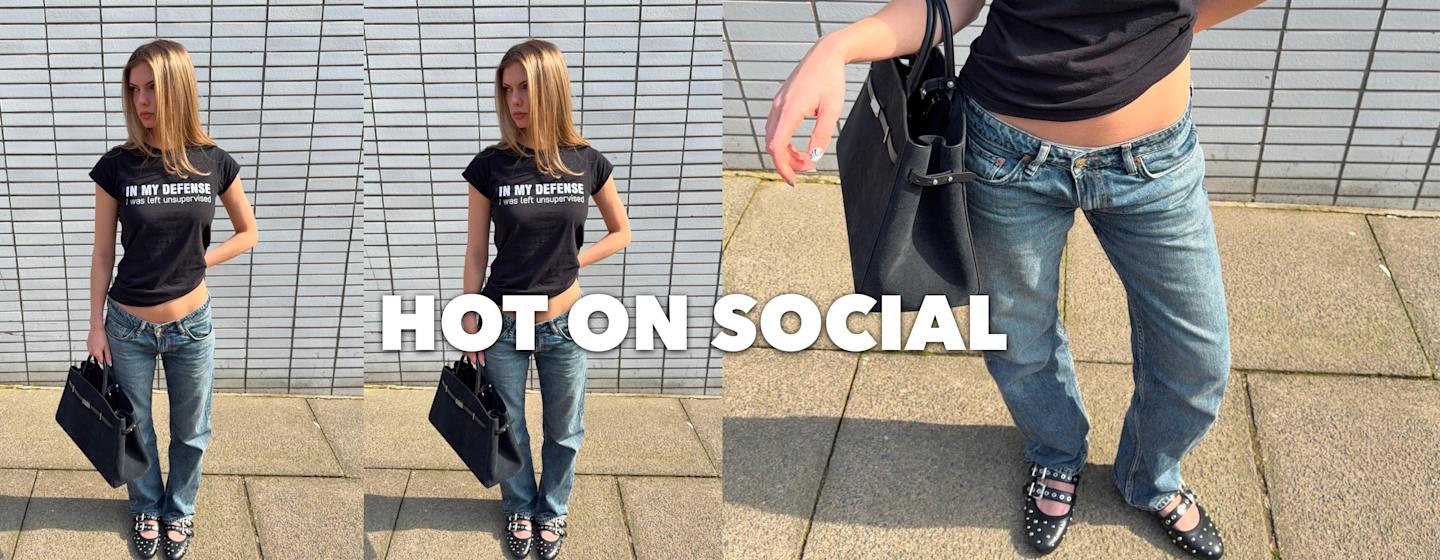 Woman in a black graphic T-shirt and blue jeans holding a black handbag, standing on a sidewalk against a tiled wall, with studded black shoes visible.