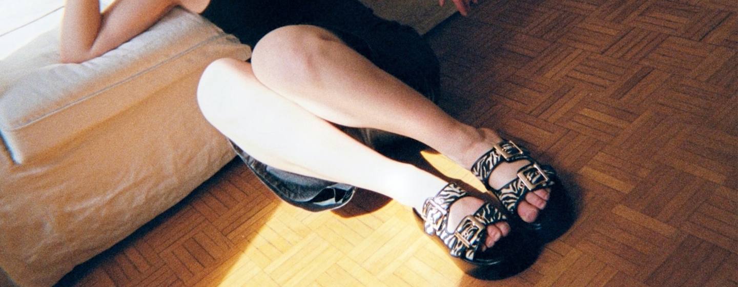 Person reclining on the floor with legs extended, wearing patterned sandals, next to a sofa in warm sunlight.