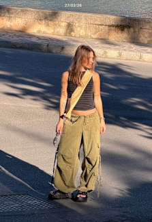 A woman in a black tank top and loose green cargo pants stands on a sunlit street near the waterfront, looking to the side.