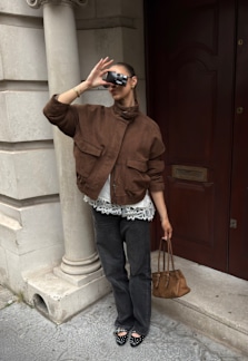 A woman in a brown jacket and dark jeans stands by a doorway, holding a phone up to her face and carrying a small handbag.