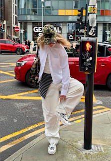 Person in a white outfit and camouflage cap lifting one foot while standing at a city crosswalk, with a red car and pedestrian signal in the background.