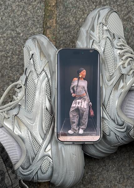 A smartphone placed between a pair of worn gray sneakers displays a photo of a woman posing in a stylish outfit, standing on a small platform.